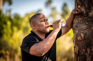 Man in black shirt playing trumpet outdoors near a tree with lush green background.