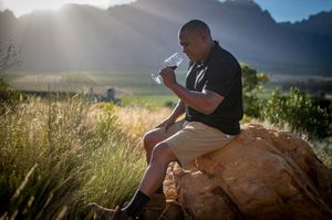 Man sitting on a rock in a scenic mountain valley, tasting wine with sunlight streaming through the background.