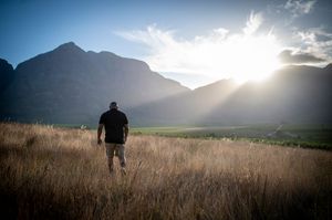 Man standing in golden grassland gazing at sunset over dramatic mountain valley landscape.