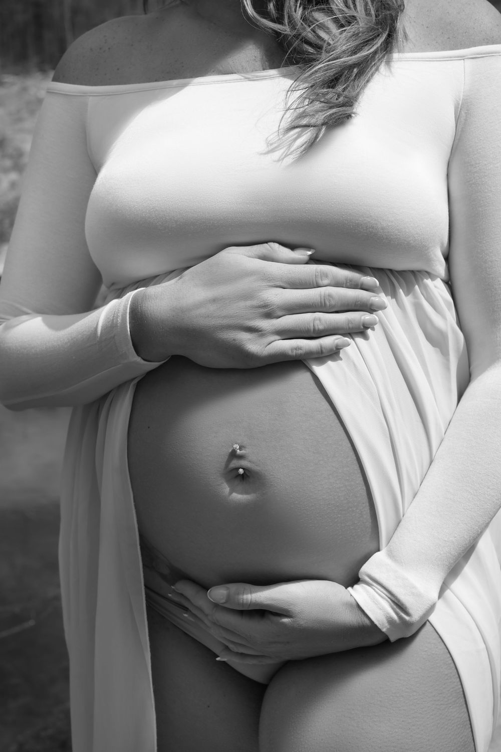 Black and white photo of a pregnant woman cradling her bare baby bump, wearing a flowing white maternity gown outdoors.