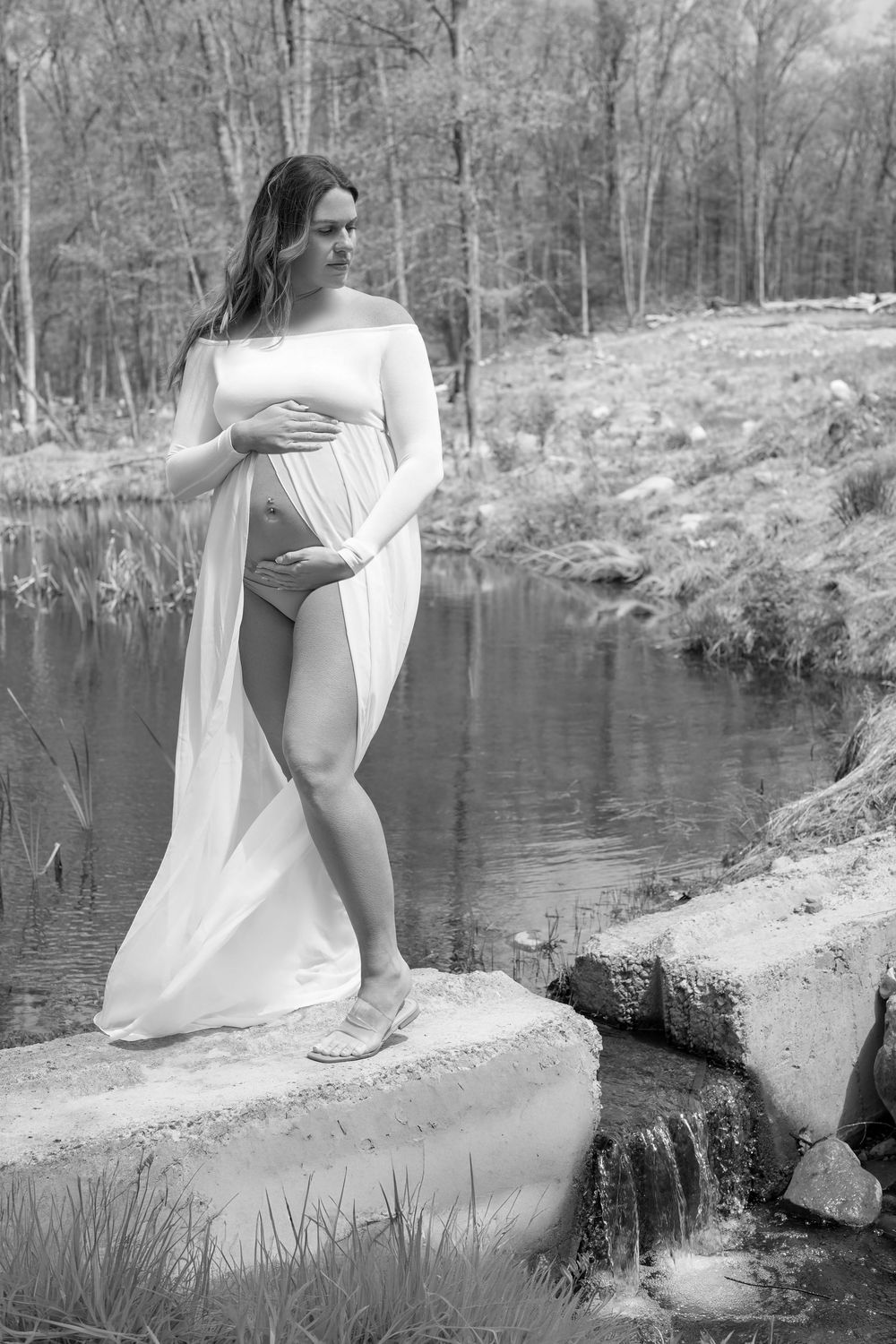 Black and white photo of pregnant woman in white draped fabric standing on rocks by a serene pond in a forest.