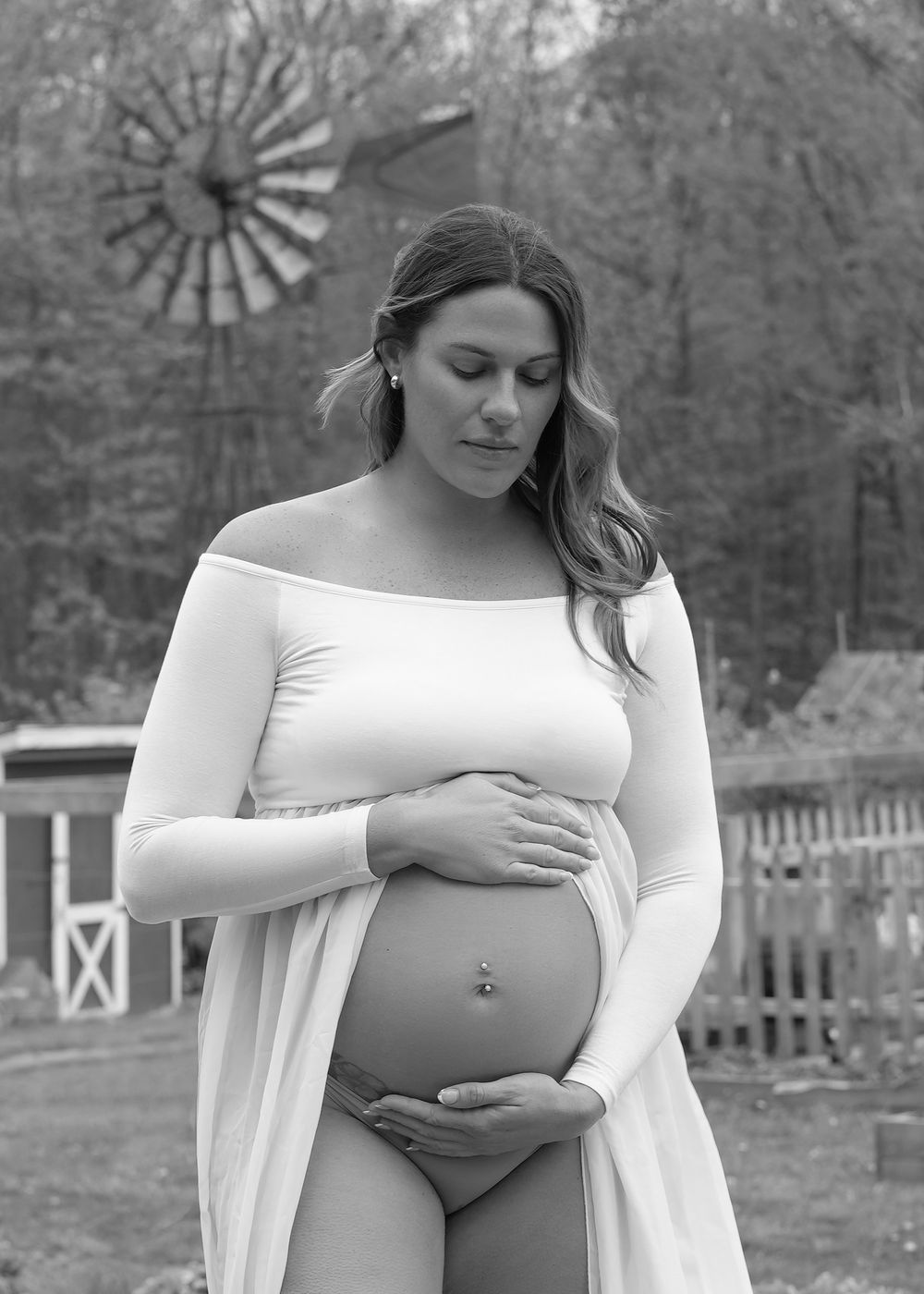 Black and white maternity photo of a pregnant woman in a white off-shoulder dress outdoors near a windmill.