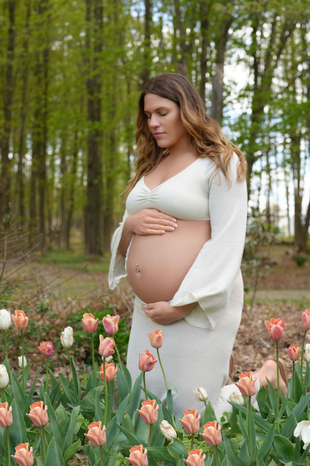 Pregnant woman in white dress cradling baby bump among pink and white tulips in a lush green forest setting.