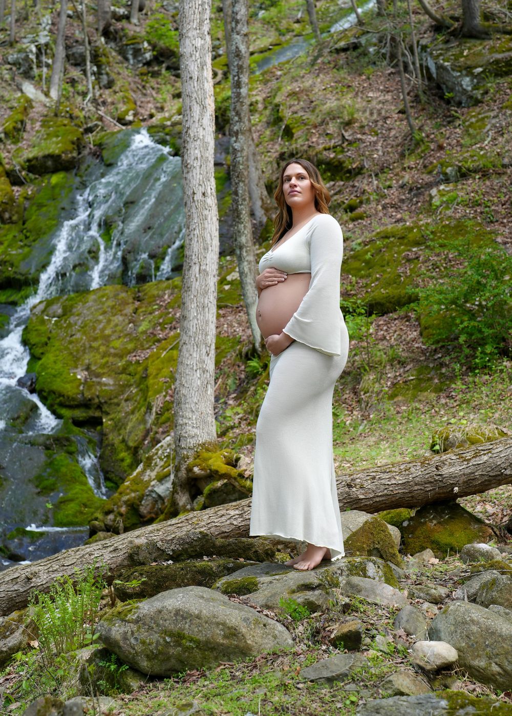 Pregnant woman in white dress poses by mossy waterfall in forest setting.