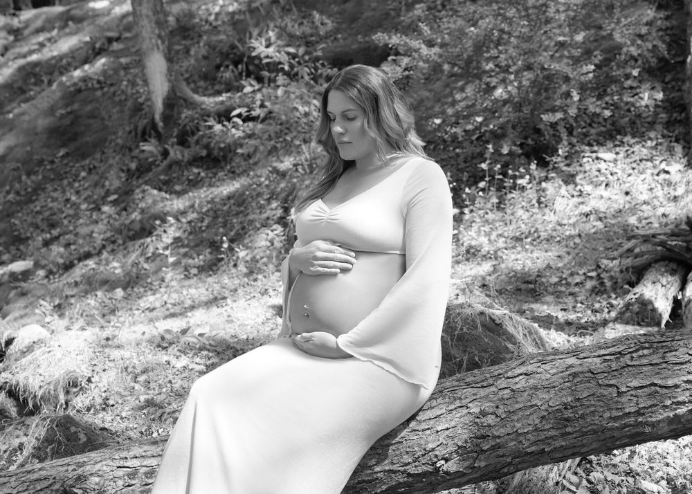 Black and white maternity photo of a pregnant woman in a white gown sitting on a log in a forest.
