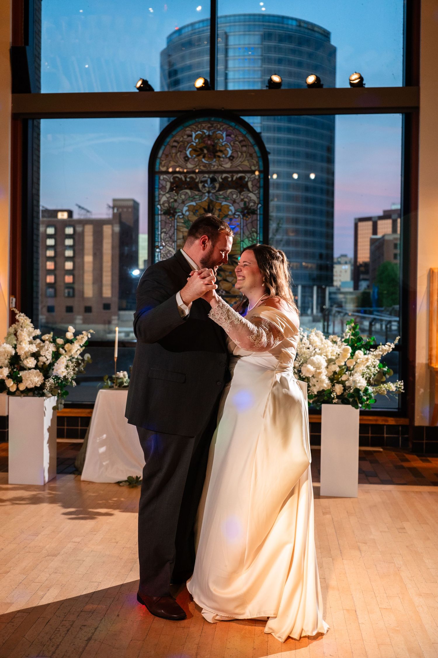Bride and groom share first dance in elegant venue with city skyline and stained glass window backdrop.