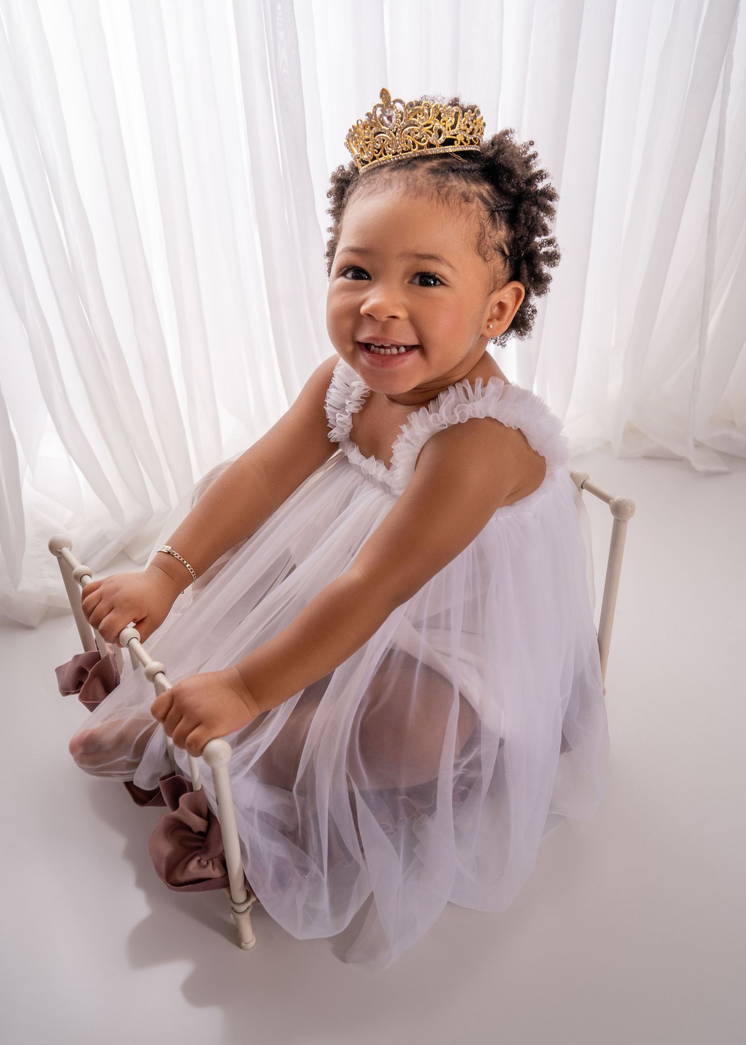 A smiling toddler girl in a white tulle dress and gold tiara sits on a small chair in a bright white studio.