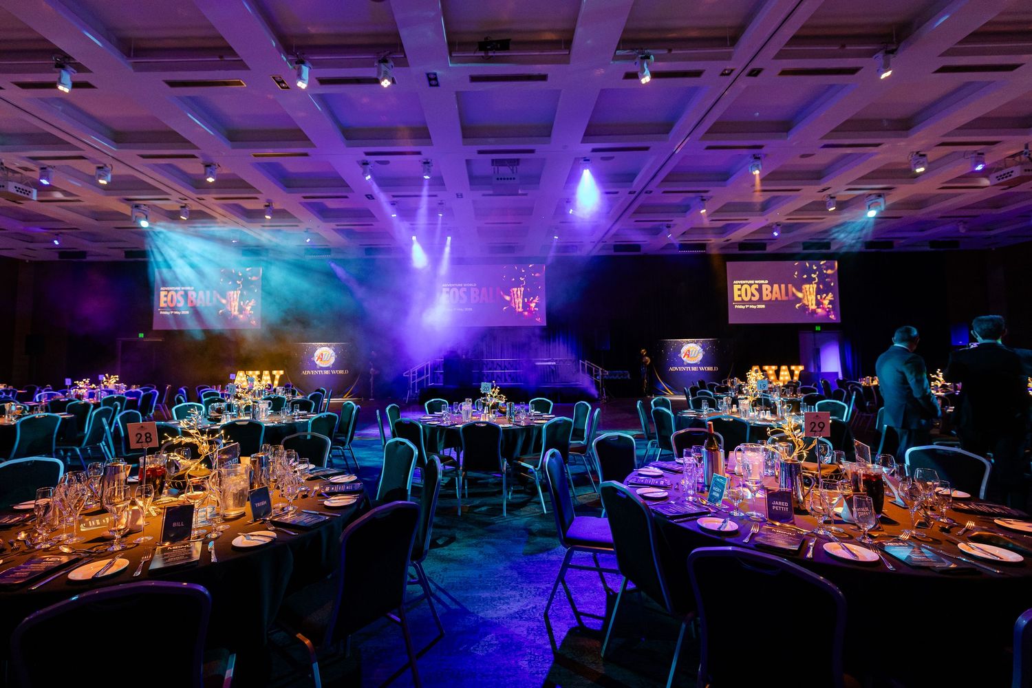 Elegant gala dinner setup with round tables, blue and purple stage lighting, and branded screens in a large event hall.