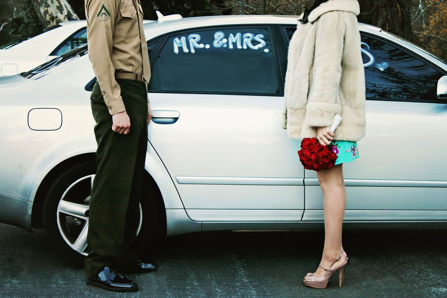 Newlywed couple stands beside a silver car marked Mr. and Mrs., the bride holding red roses and the groom in a military uniform.