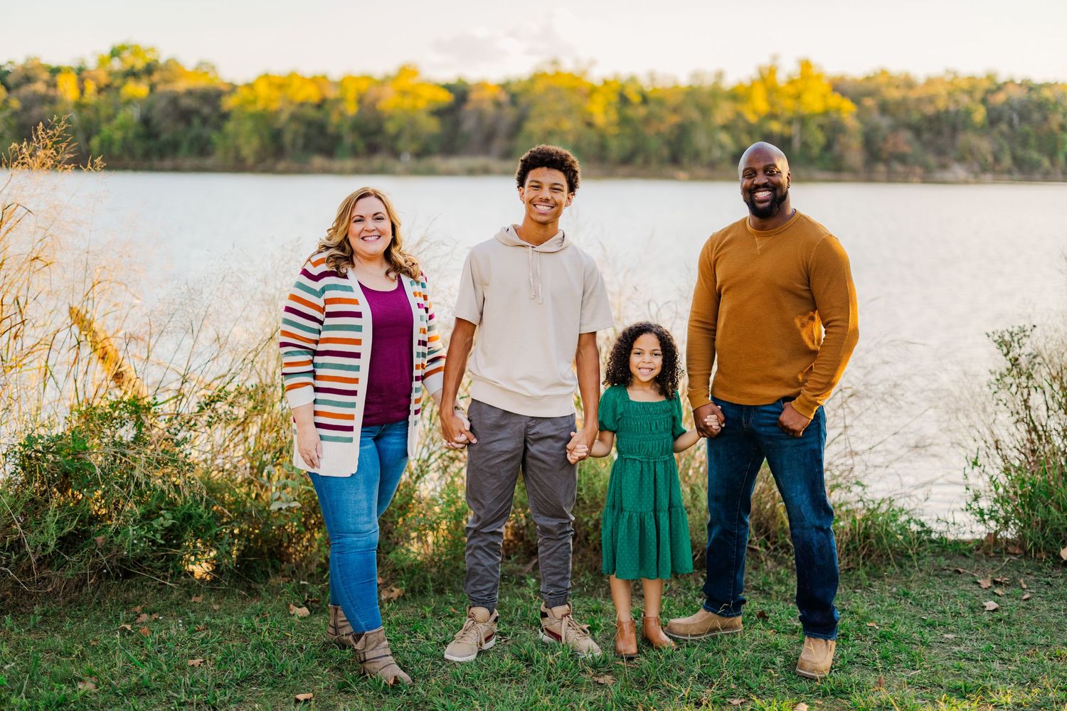 Family of four smiling outdoors by a lake in autumn, wearing colorful fall outfits on green grass.