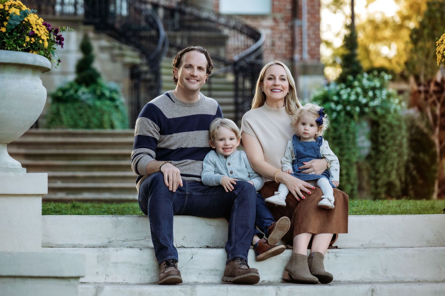 Happy family of four sitting on outdoor steps, parents with two young children, smiling in a garden setting.