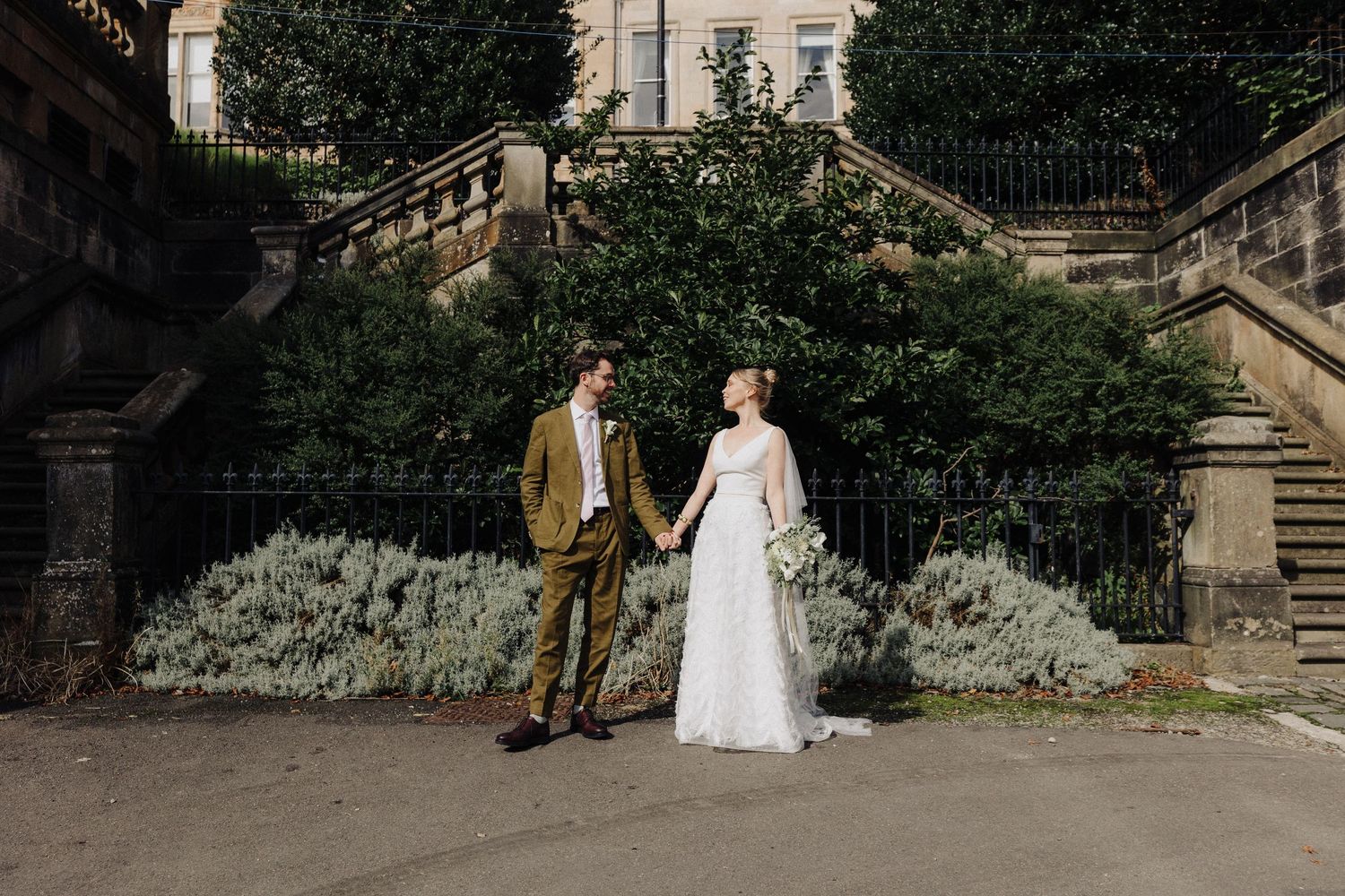 Bride and groom hold hands in front of grand stone staircase and lush garden greenery.