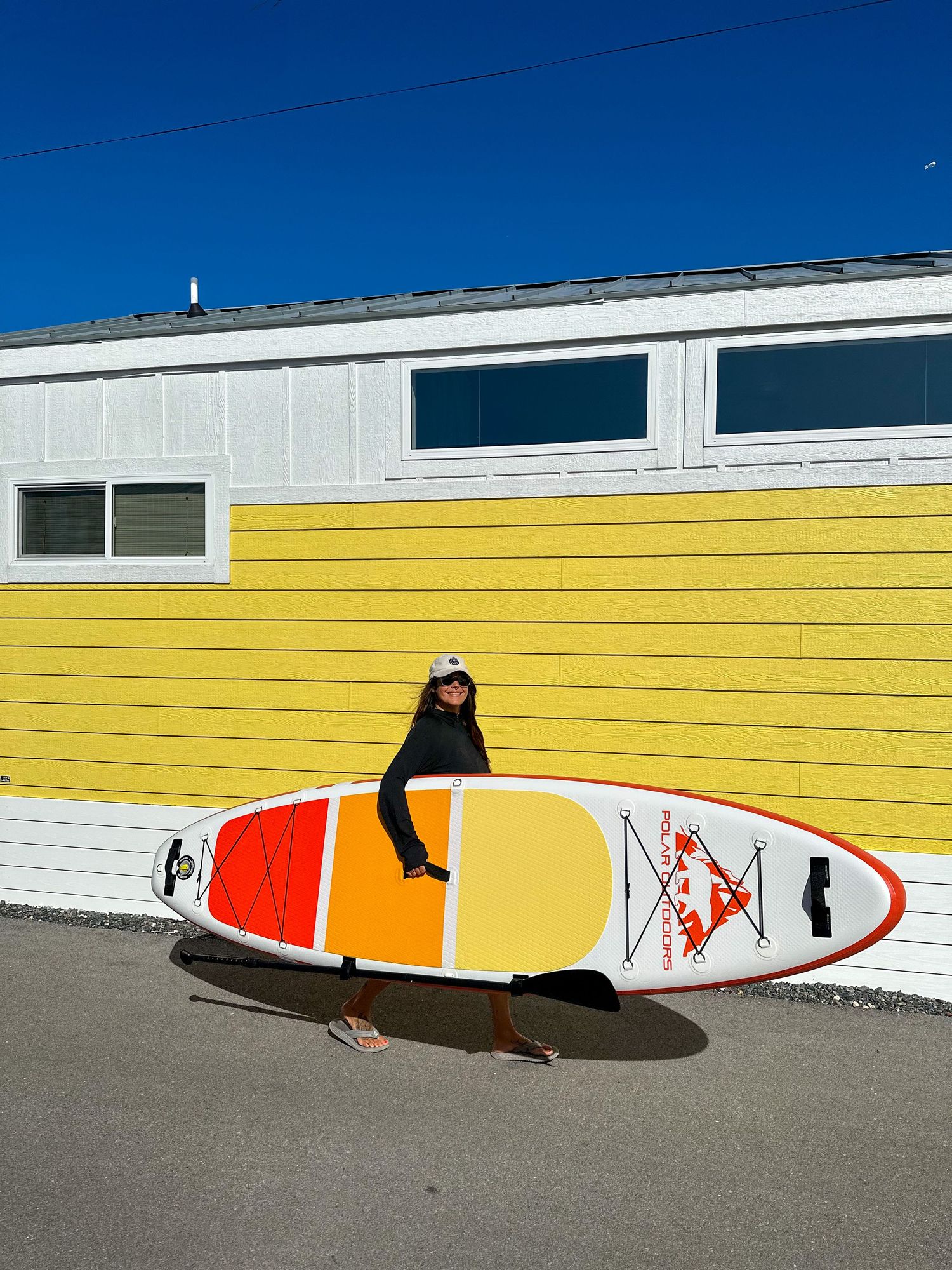 Woman holding a large orange and yellow inflatable paddleboard in front of a yellow building outdoors.