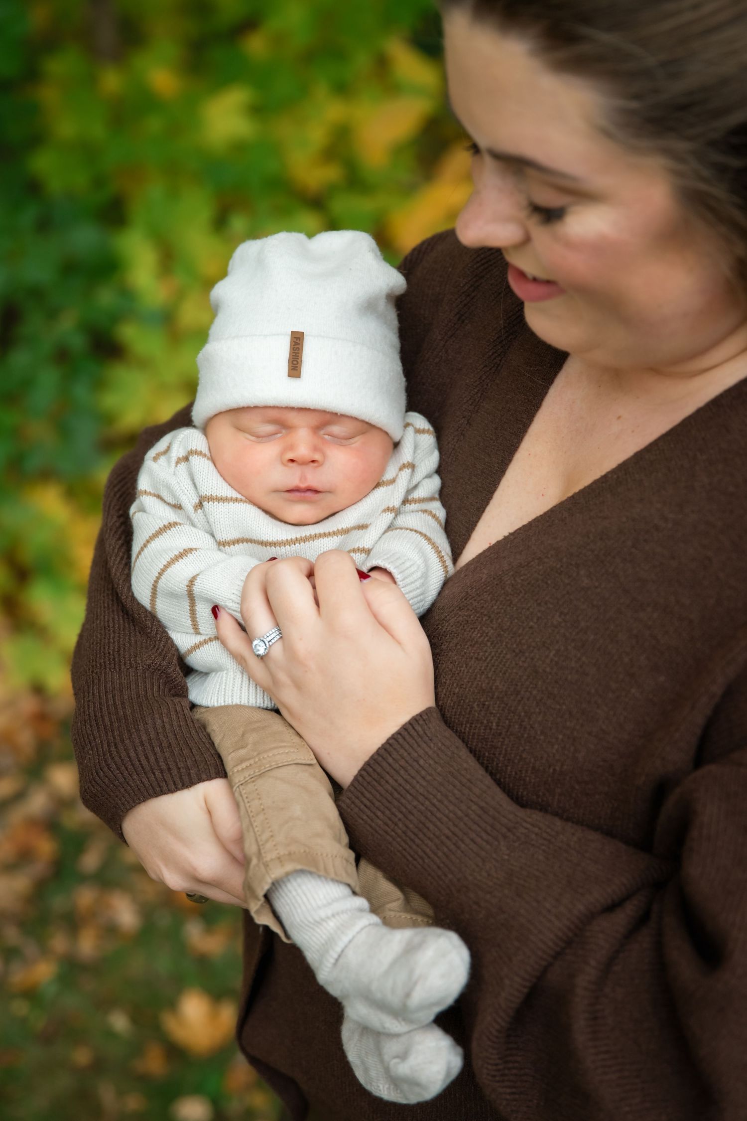 Mother in brown sweater cradling sleeping newborn baby in white hat outdoors in autumn park.
