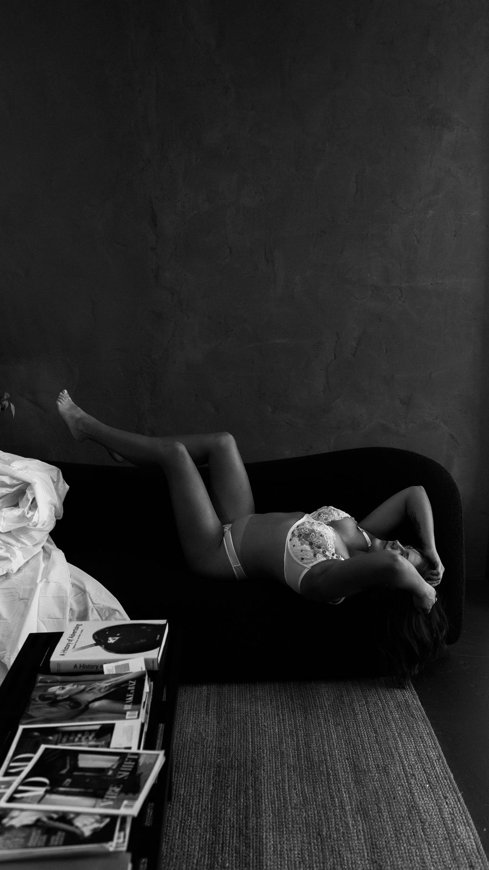 Black and white boudoir photo of woman in white lingerie lying on dark sofa with magazines nearby.