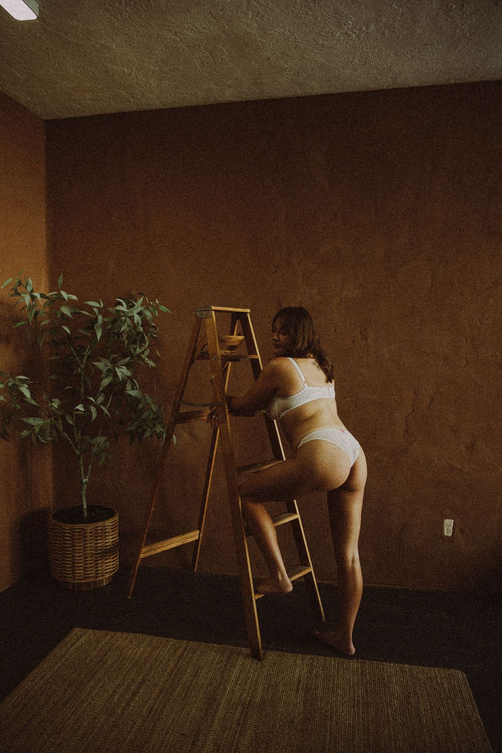 Woman in white lingerie climbing a wooden ladder in a warmly lit room with a potted plant.