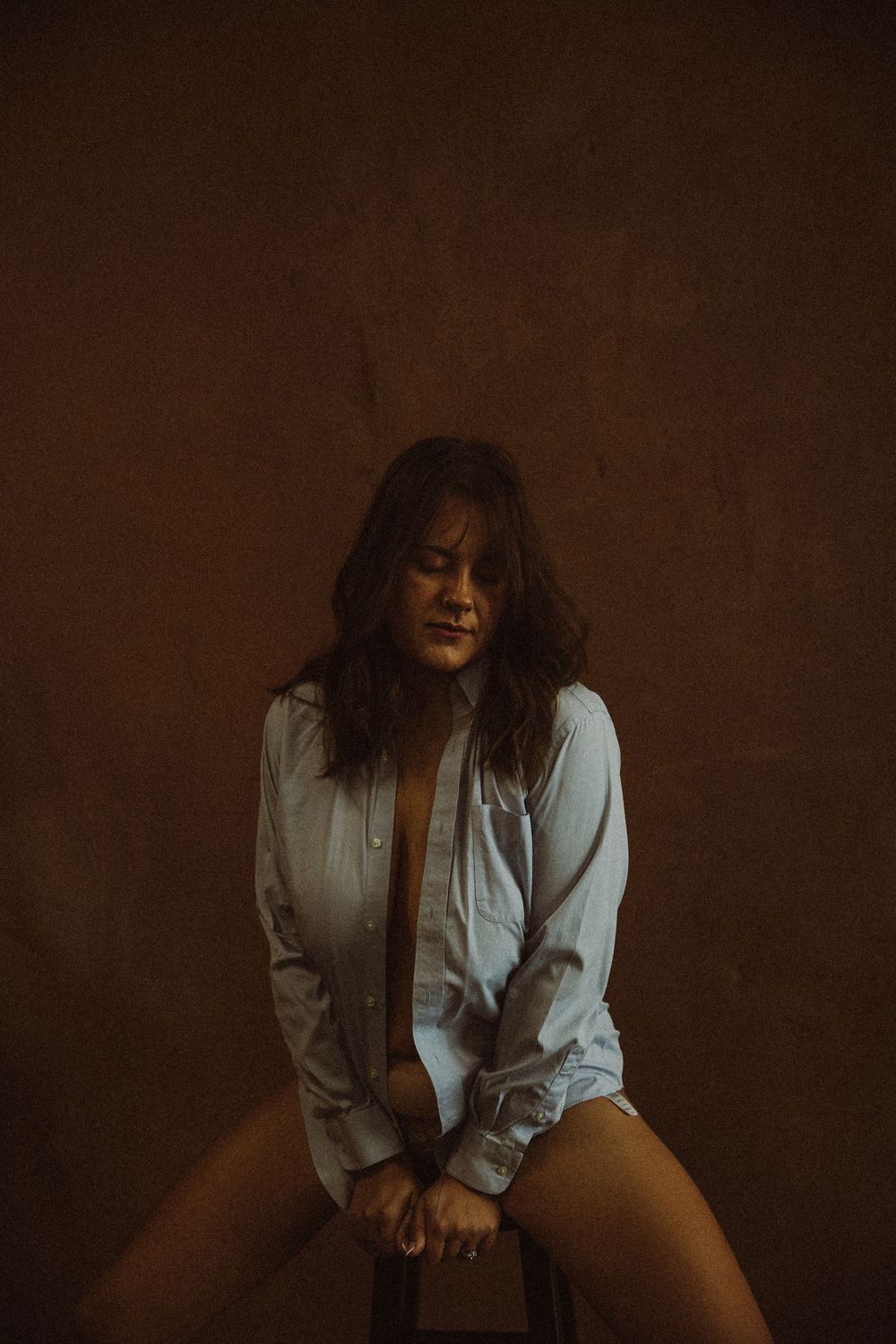 Woman with long hair sitting on a stool in an open white shirt in moody warm lighting.
