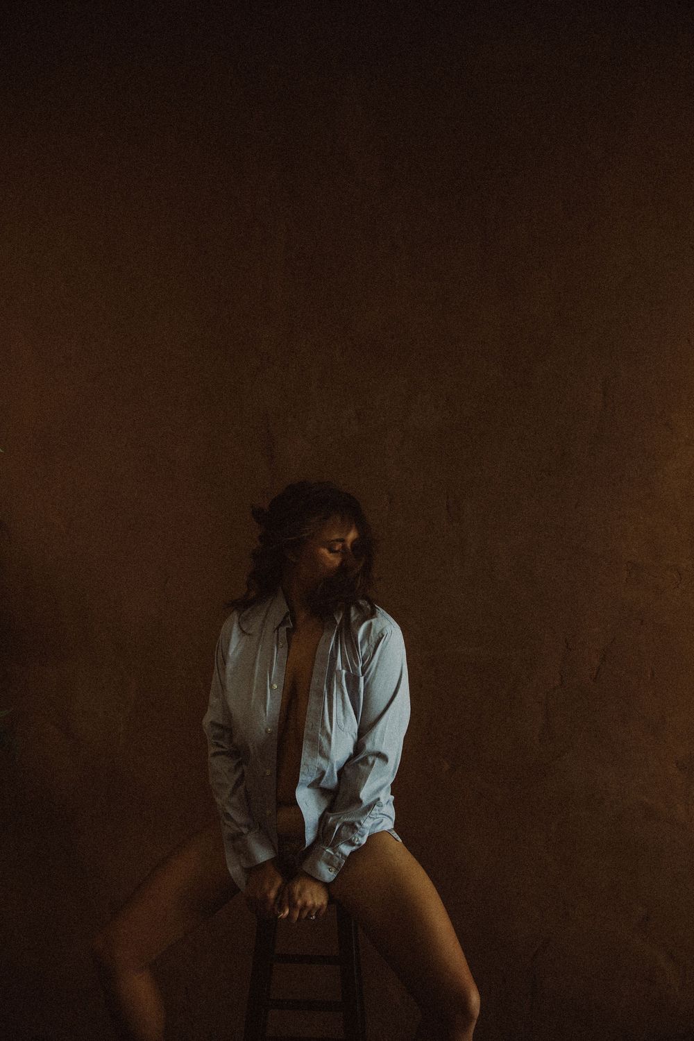 Woman in an open white shirt seated on a stool against a dark warm-toned background.