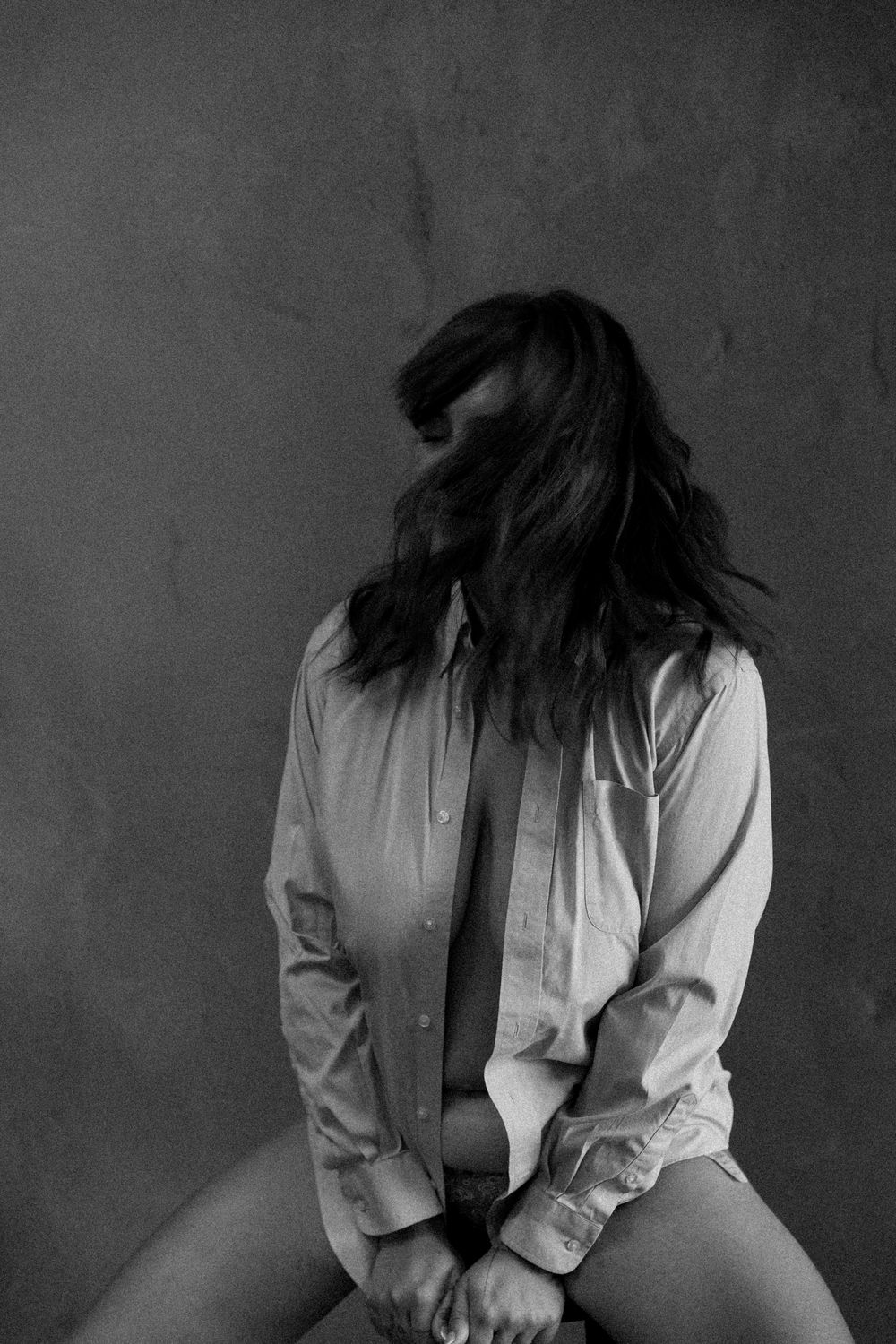 Black and white photo of a woman in an open shirt with hair covering her face on a stool.