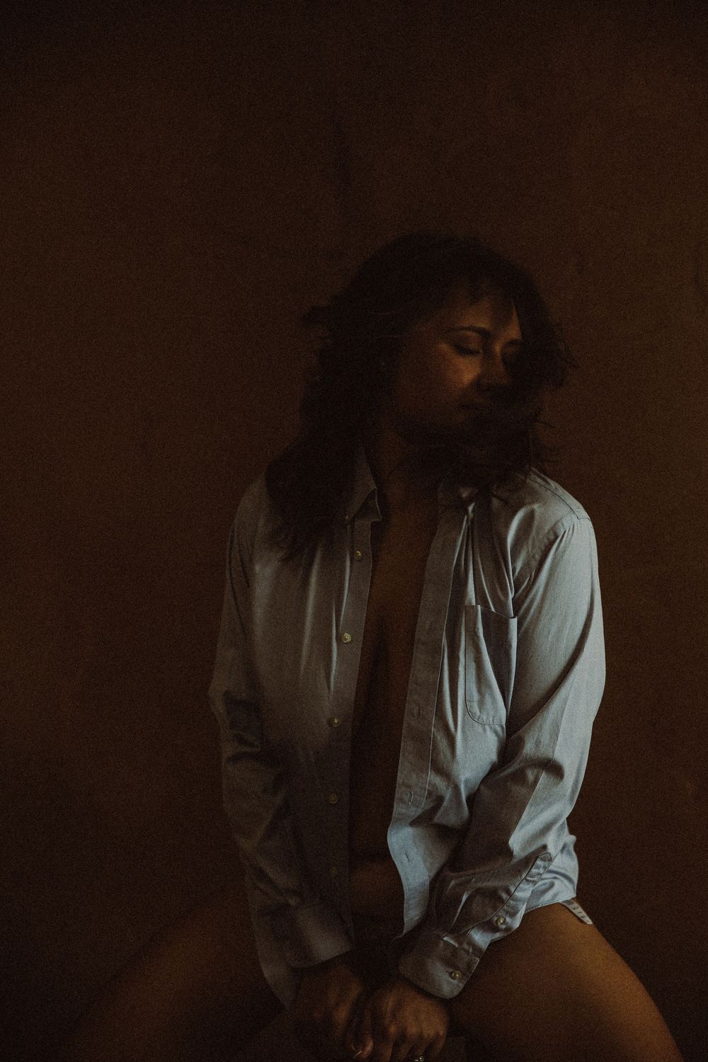 Woman in an unbuttoned white shirt sitting with eyes closed in dramatic warm studio light.