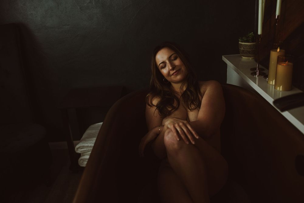 Woman relaxing in a dark, candlelit bathtub with a serene expression in a moody bathroom setting.