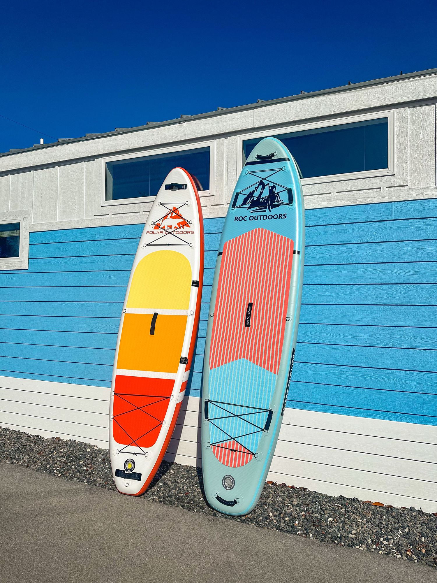 Two colorful inflatable ROC Outdoors stand-up paddleboards leaning against a bright blue and white building.