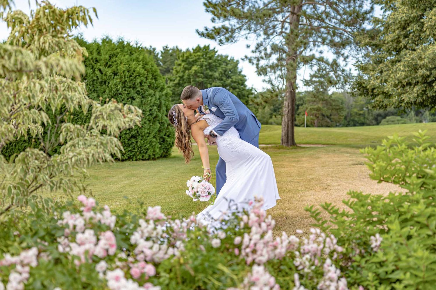 Groom dips bride for romantic kiss among pink blooming flowers in lush garden setting. Stonebridge Country Club Goffstown NH