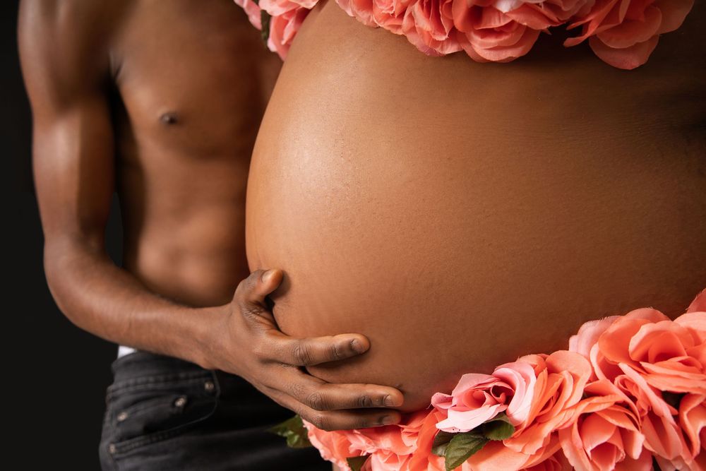 Pregnant woman's bare belly surrounded by pink roses with partner's hand gently holding her side.