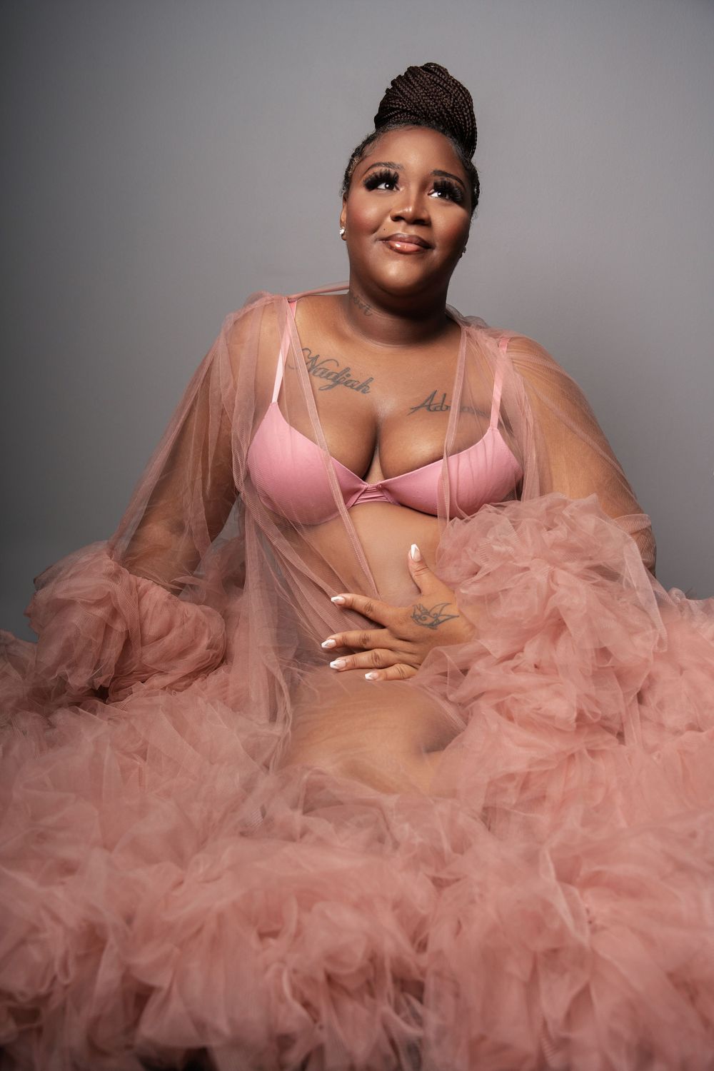 Pregnant woman in pink bra and tulle robe sitting and smiling with tattoos visible on chest.