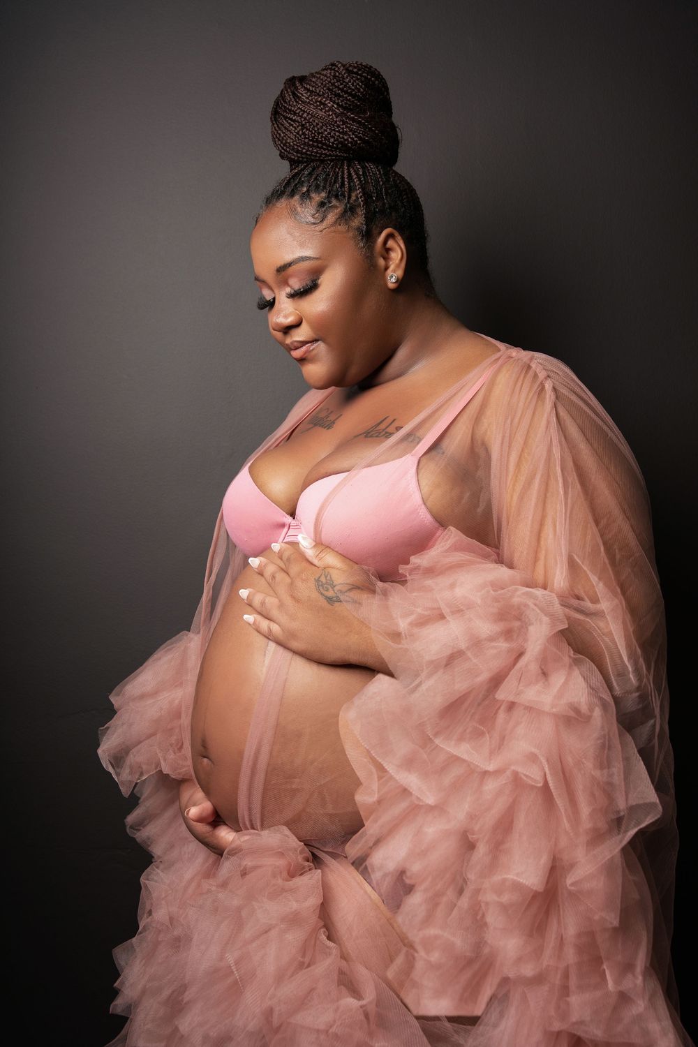 Pregnant woman in pink bra and ruffled tulle robe cradling her baby bump against dark background.