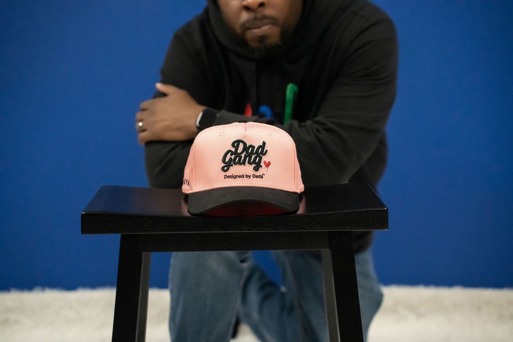 Man in black hoodie posing with a pink Dad Gang cap displayed on a black stool.