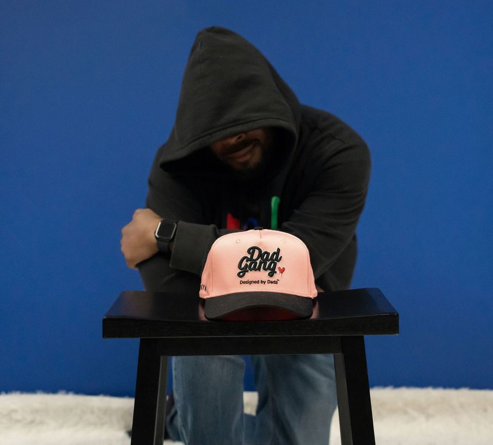 Person in black hoodie leaning over a pink Dad Gang branded cap on a black stool.