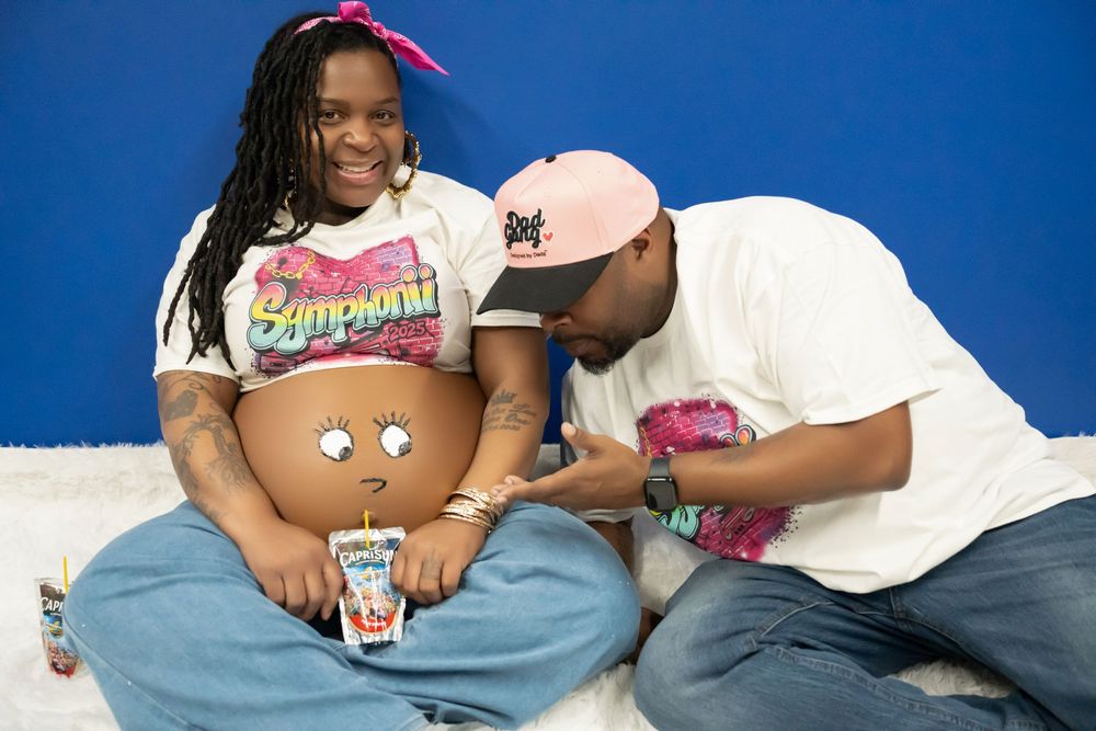 Man painting cartoon face on pregnant woman's baby bump during creative maternity shoot.