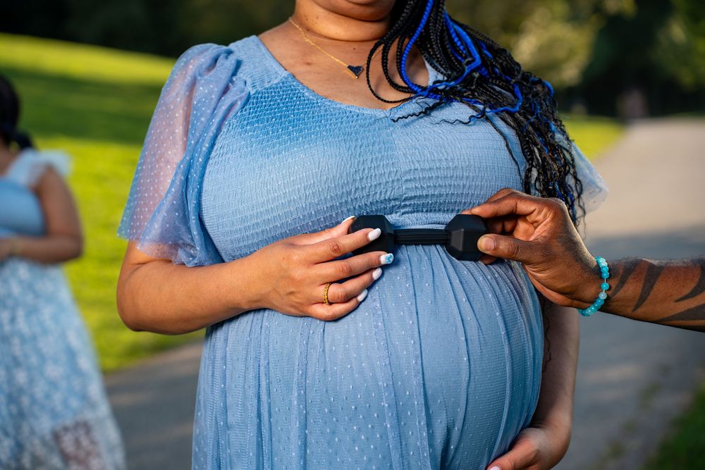 Pregnant woman in blue dress holds her baby bump while someone places a small bow on her belly outdoors.