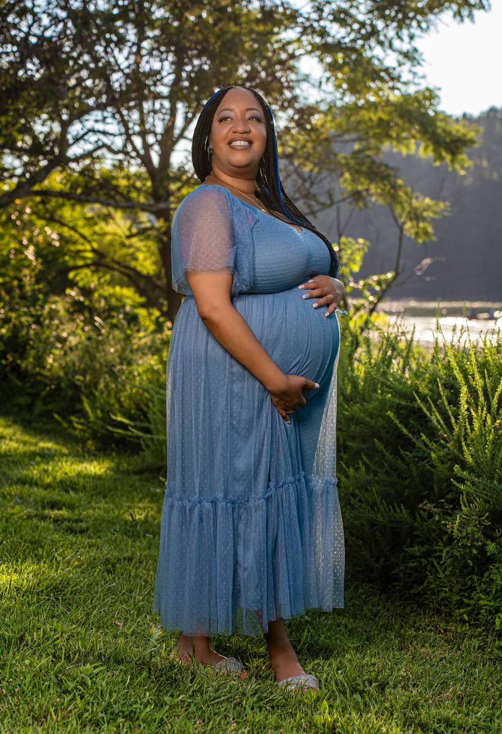 Smiling pregnant woman in blue tulle maternity dress cradles her baby bump in a lush green outdoor setting.