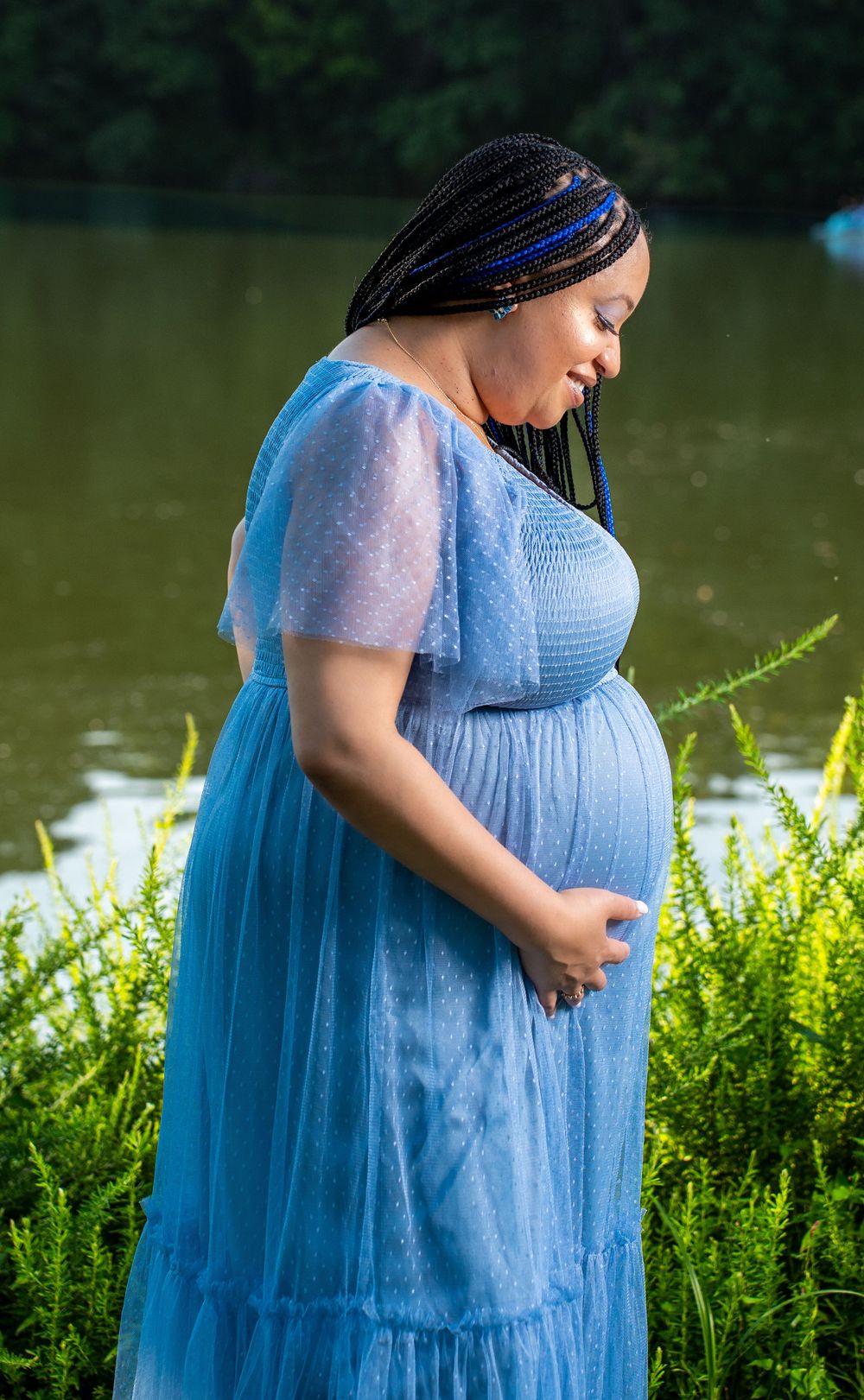 Pregnant woman in blue dress gently holds her baby bump while standing near a peaceful lakeside with greenery.