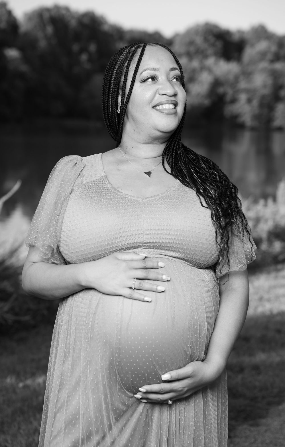 Black and white maternity portrait of a pregnant woman with braids holding her belly and gazing upward outdoors.