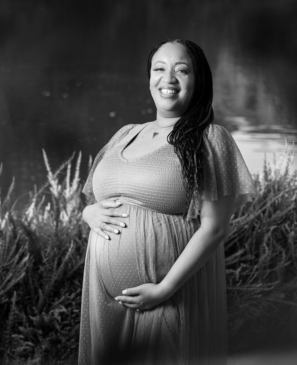 Black and white portrait of a smiling pregnant woman in a flowing dress cradling her baby bump outdoors.
