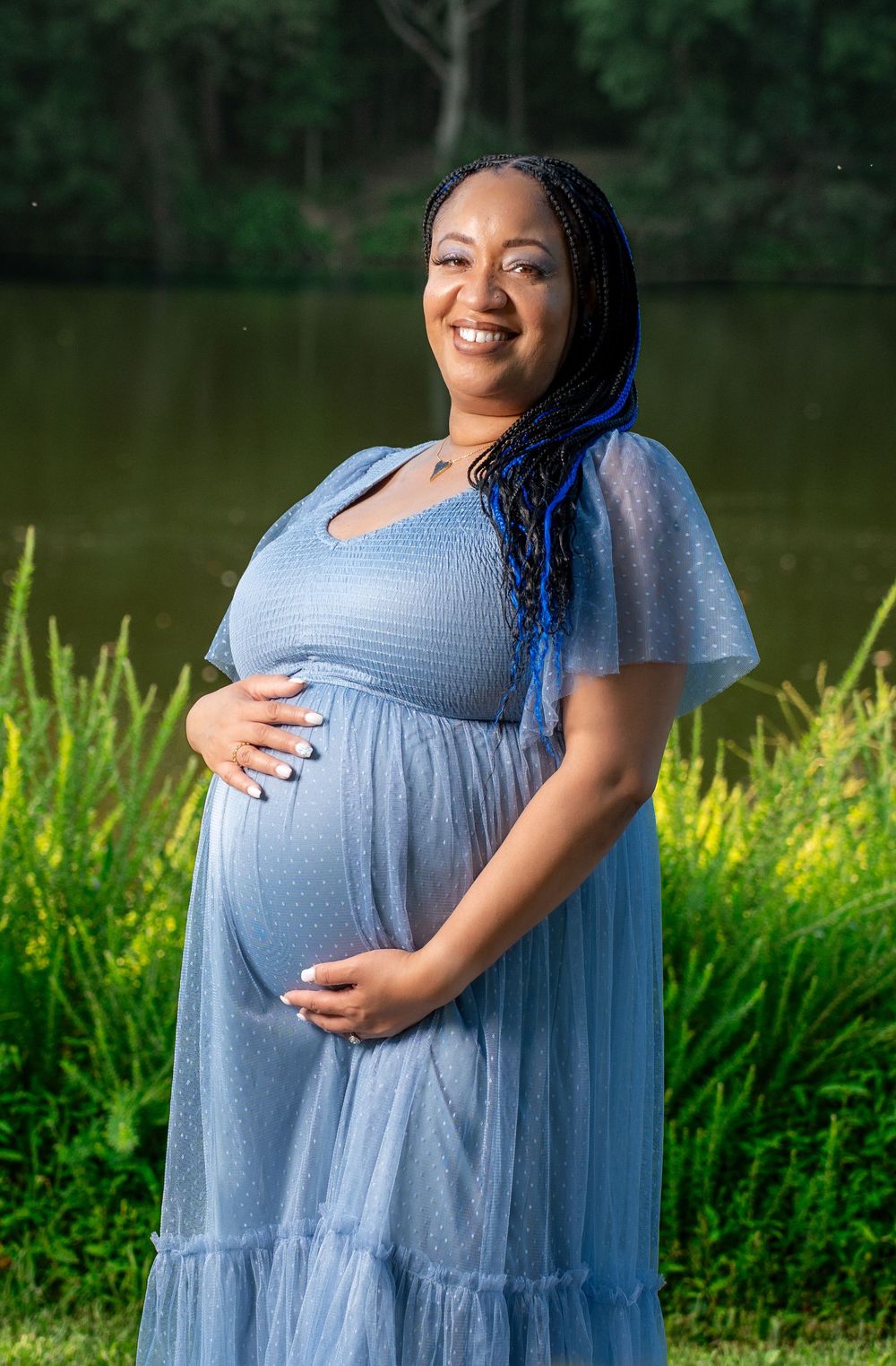 Smiling pregnant woman with blue braids in a blue dress cradles her baby bump near a serene lake.