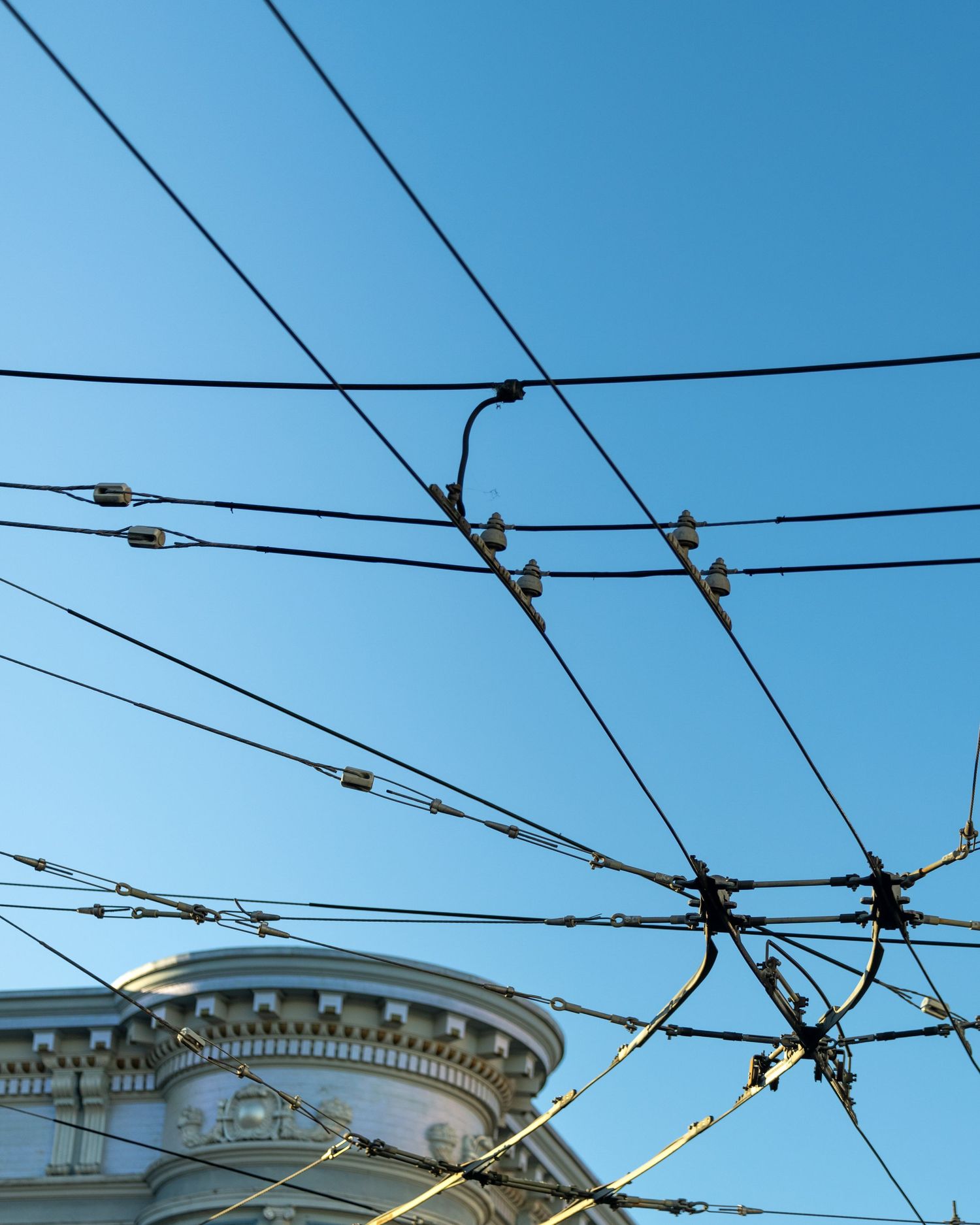 Overhead trolley wires and insulators crisscross against a clear blue sky above a classical building facade.