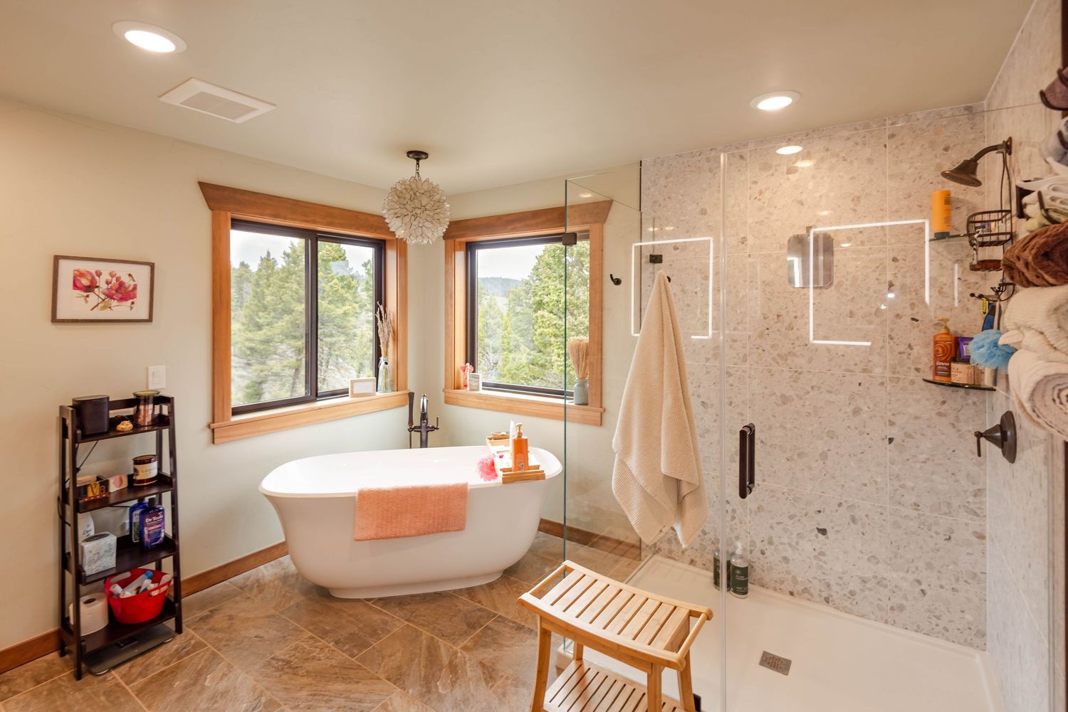 Luxurious bathroom featuring a freestanding soaking tub, walk-in tiled shower, wood bench, and chandelier with forest views.