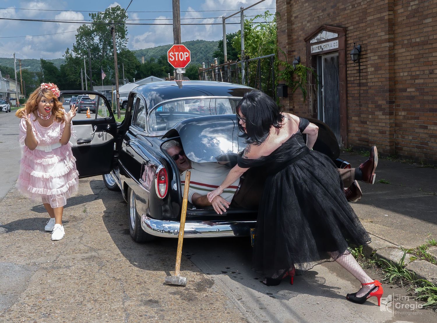 Two women in vintage 1950s attire pose dramatically near a classic black car on a street with a stop sign.