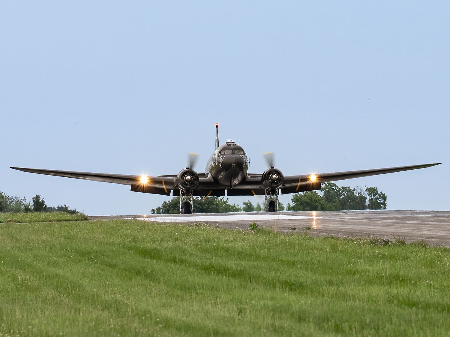 Vintage twin-engine military aircraft landing on a grass airstrip with landing lights on during an airshow.