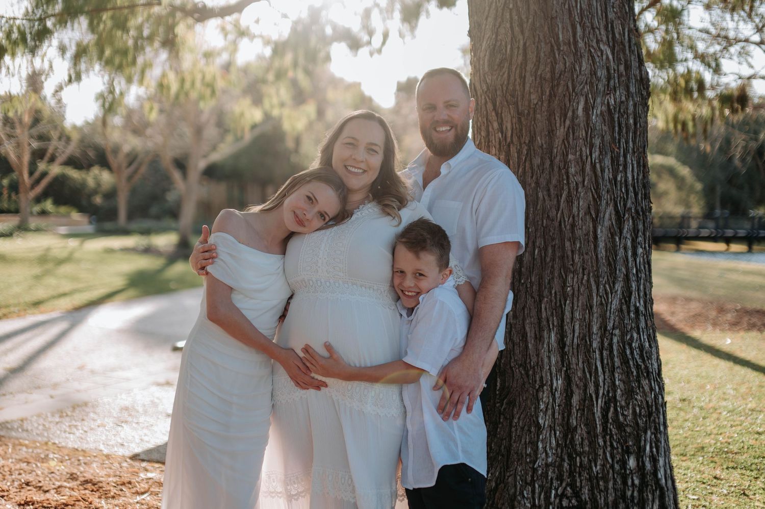 Pregnant woman in white dress poses with family near tree in sunny park, maternity photo session outdoors.