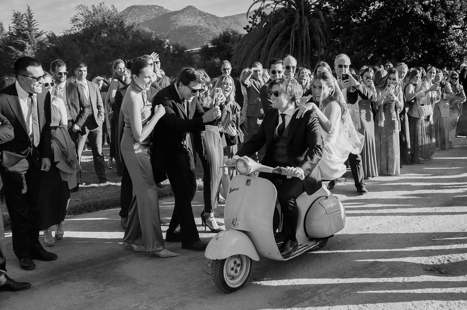 Two brides in white wedding dresses ride in a classic red convertible car through a scenic field.