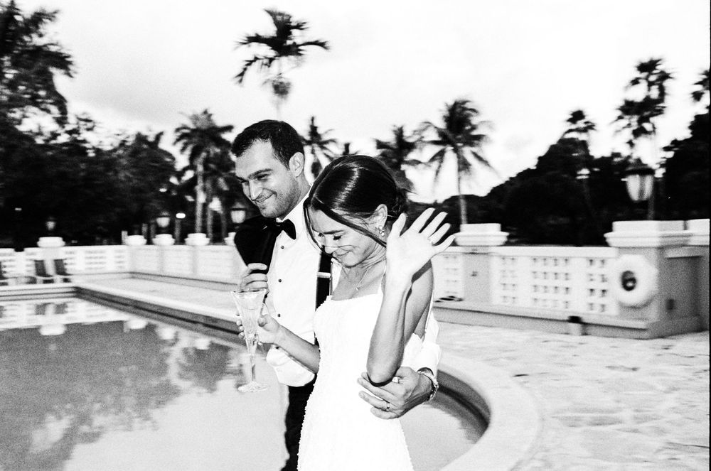 Black and white photo of a couple embracing near a pool with palm trees at a tropical wedding venue.