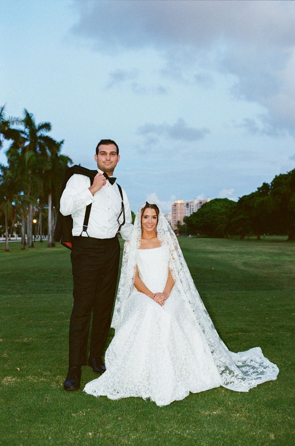 Bride in lace gown with cathedral veil stands with groom in black tuxedo on green lawn at dusk.