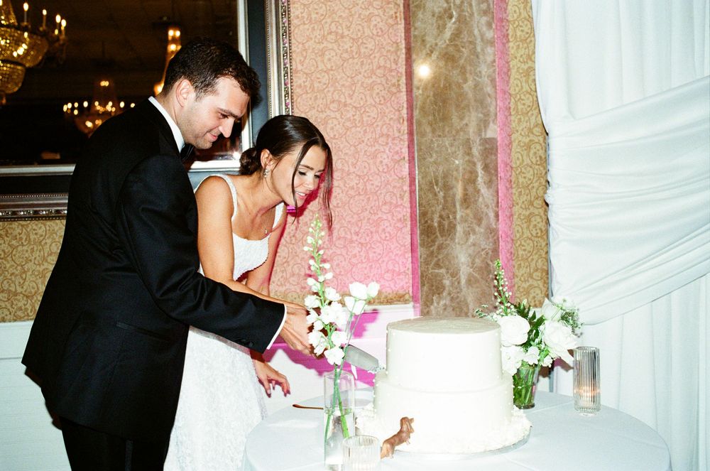 Bride and groom cutting white wedding cake together at elegant reception venue with floral arrangements.