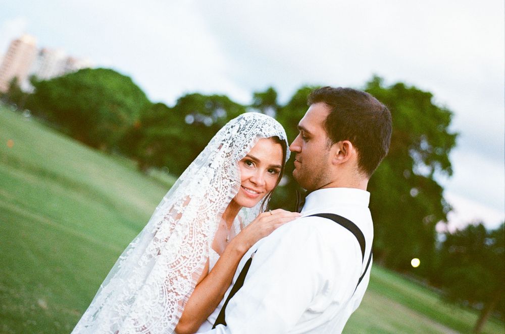 Bride in lace veil and groom in white shirt embrace outdoors on green lawn at elegant wedding.