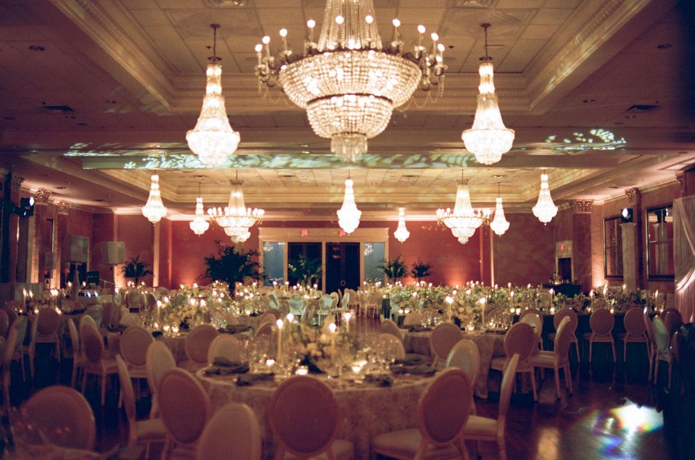 Elegant wedding reception ballroom with crystal chandeliers, round tables, white chairs, and warm pink ambient lighting.